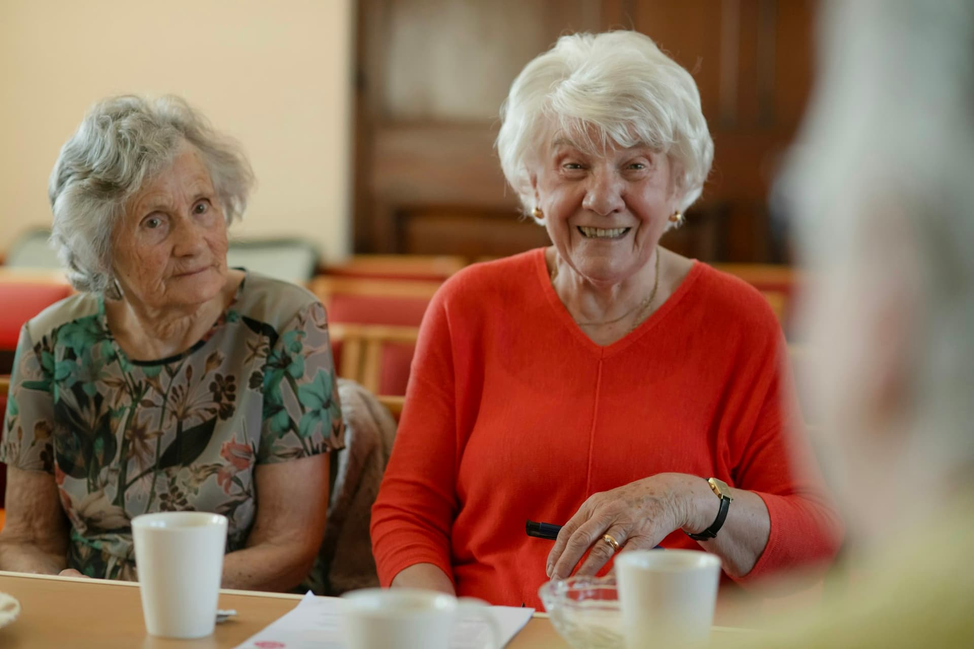 Two older women laughing together over a cup of tea at a kitchen table