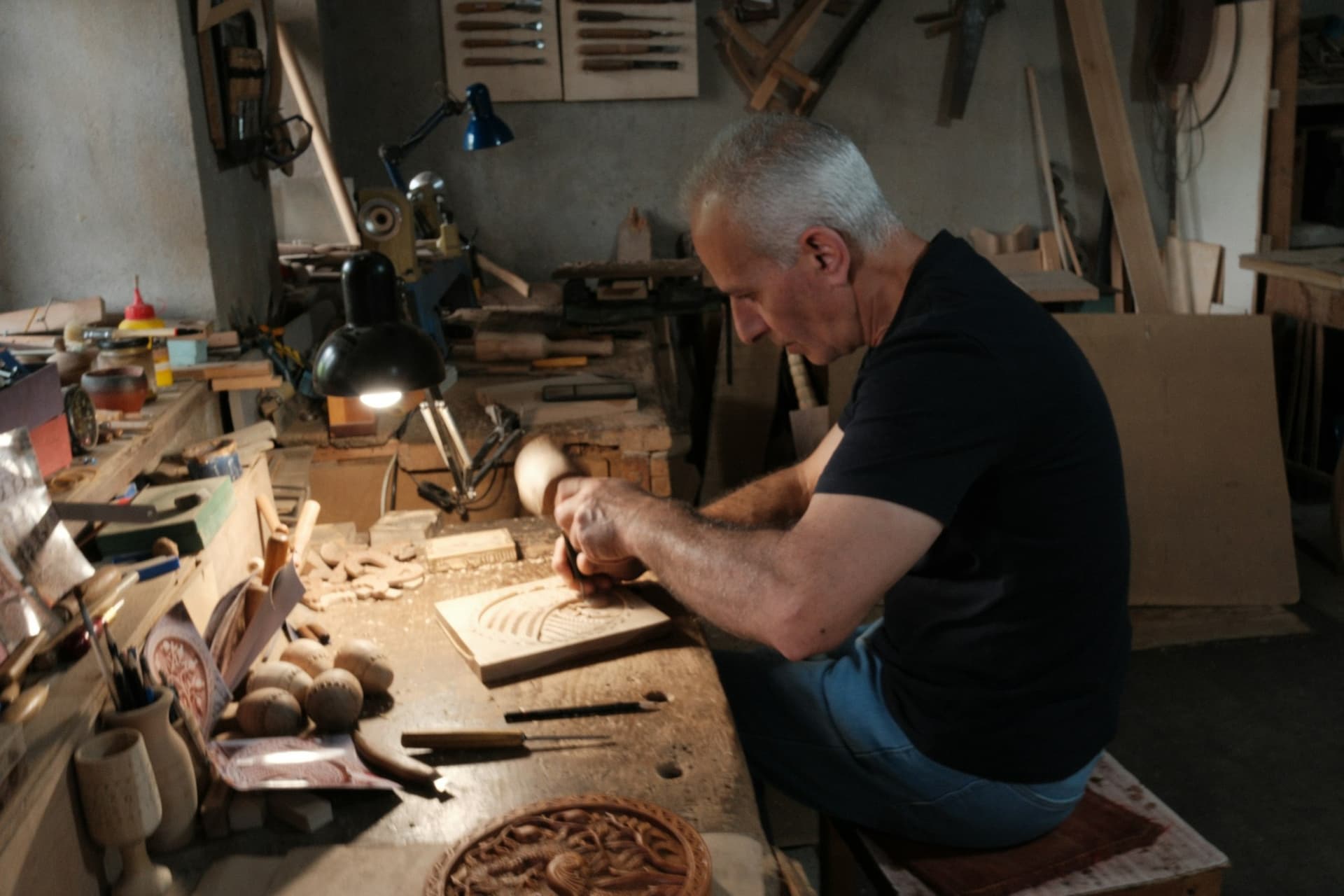 An older man carving wood at a workbench in a sunlit workshop