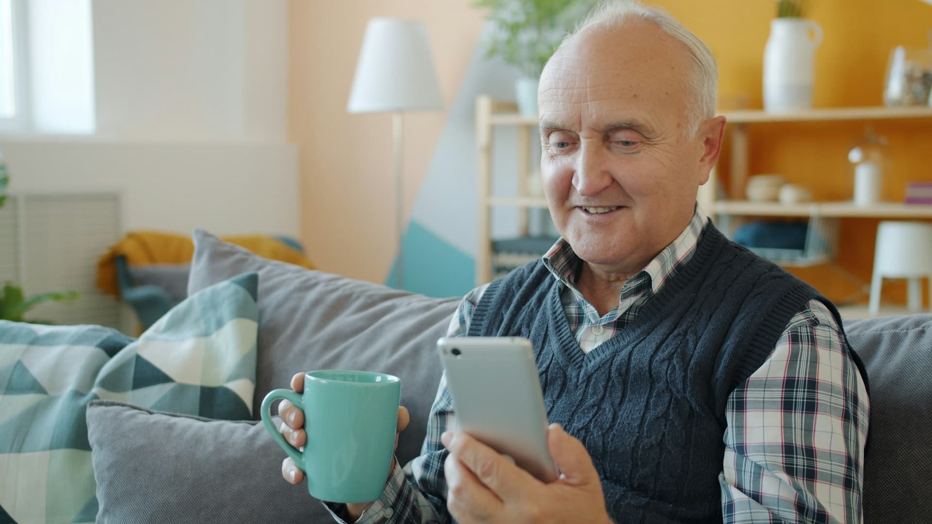A smiling older man on his sofa holding a mug and reading his phone